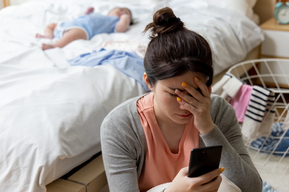 Woman looks at her smartphone with her hand on her forehead as a baby lies on the bed behind her.