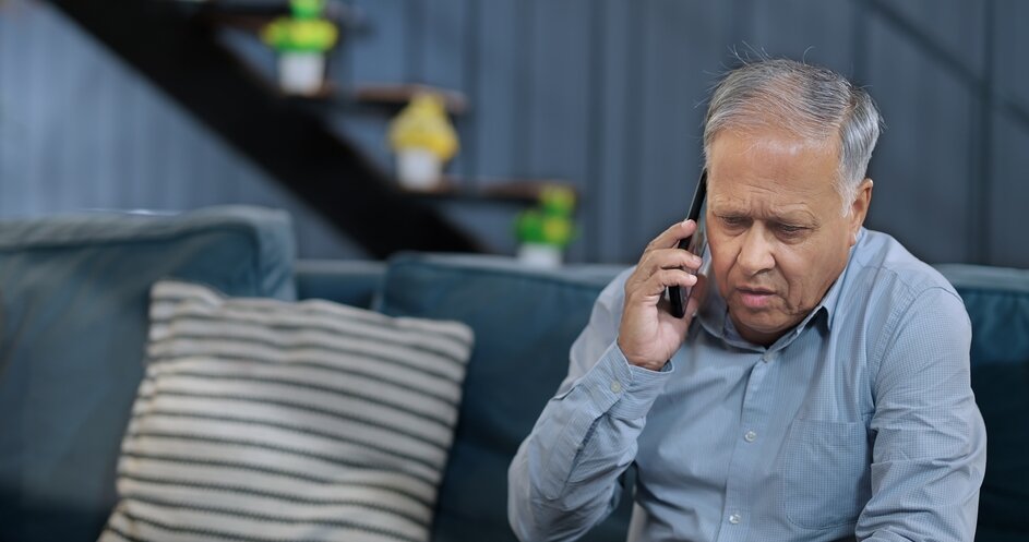Older man talking on a smartphone while seated on a couch at home.