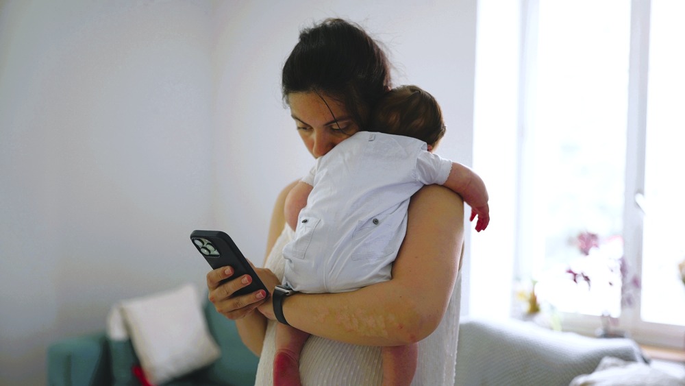 Woman checks her smartphone while holding a baby over her shoulder.