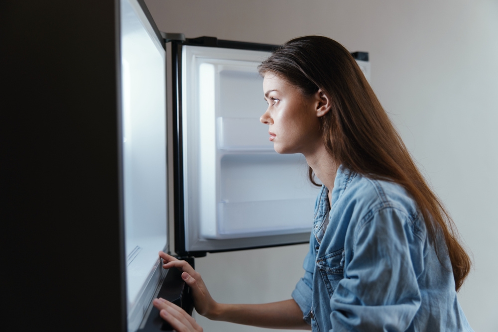Woman looks inside an open, nearly empty refrigerator.
