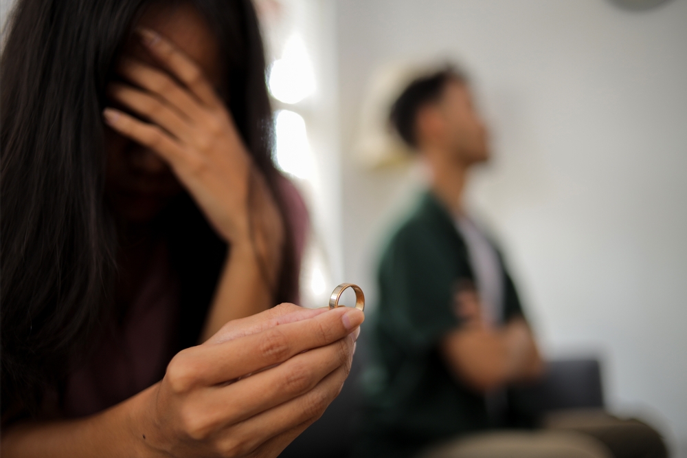 Woman holds a wedding ring as a man stands blurred in the background with his arms crossed.