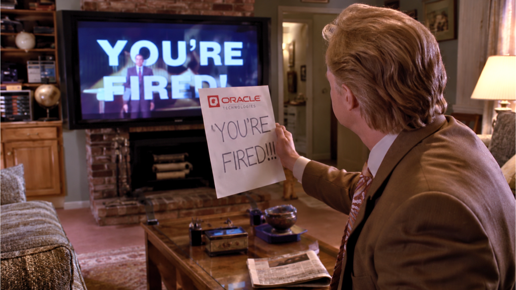 A man in a suit holding a piece of paper that reads "YOU'RE FIRED!!!" in front of a TV screen displaying the same words.