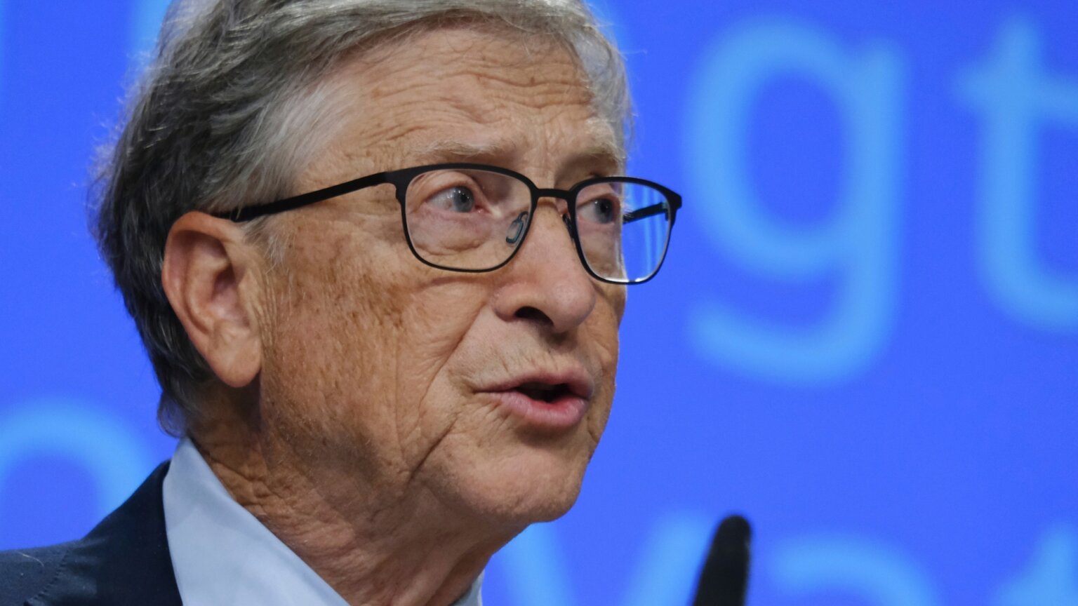 Bill Gates speaks during an event, wearing black-framed glasses against a blue background.