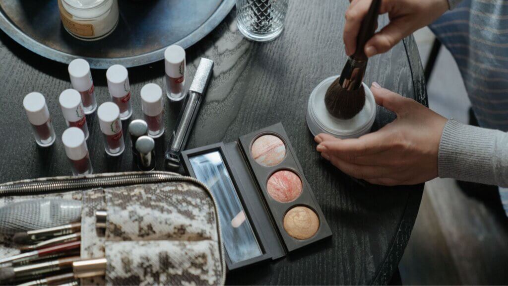 Person dipping a makeup brush into powder beside assorted cosmetics.