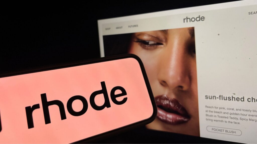 Rhode beauty website displayed behind a smartphone showing the Rhode logo.