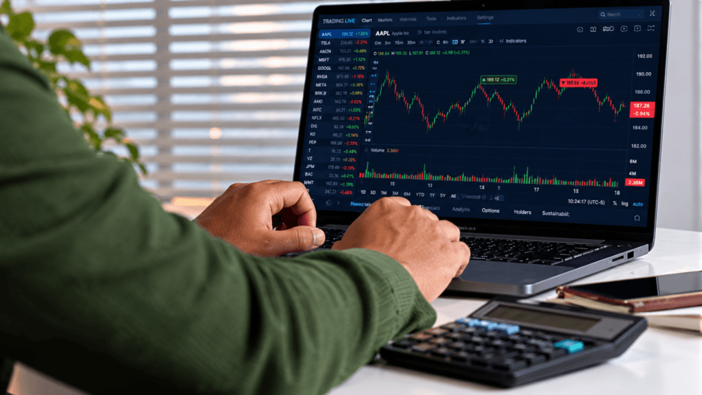 A person using a laptop to monitor stock market candlestick charts with a calculator sitting on the desk.