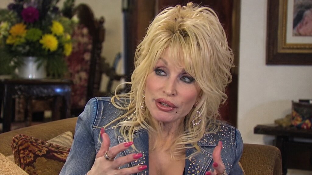 Dolly Parton gestures while speaking during an interview, wearing a rhinestone-studded denim jacket and large earrings.
