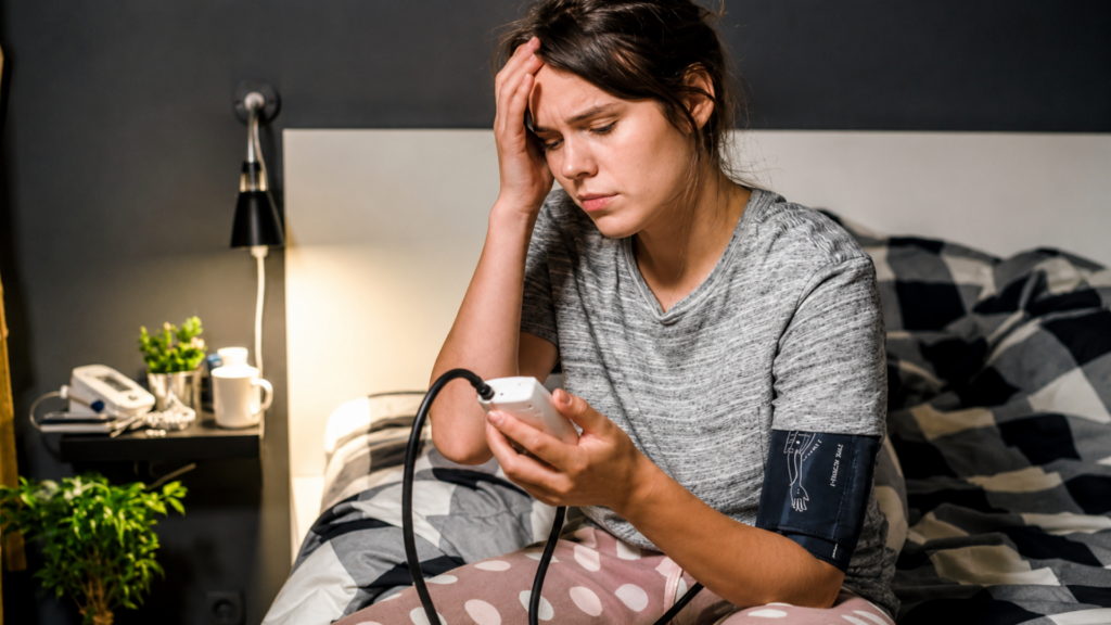 Woman sitting in bed checking her blood pressure with an electronic cuff while touching her head.