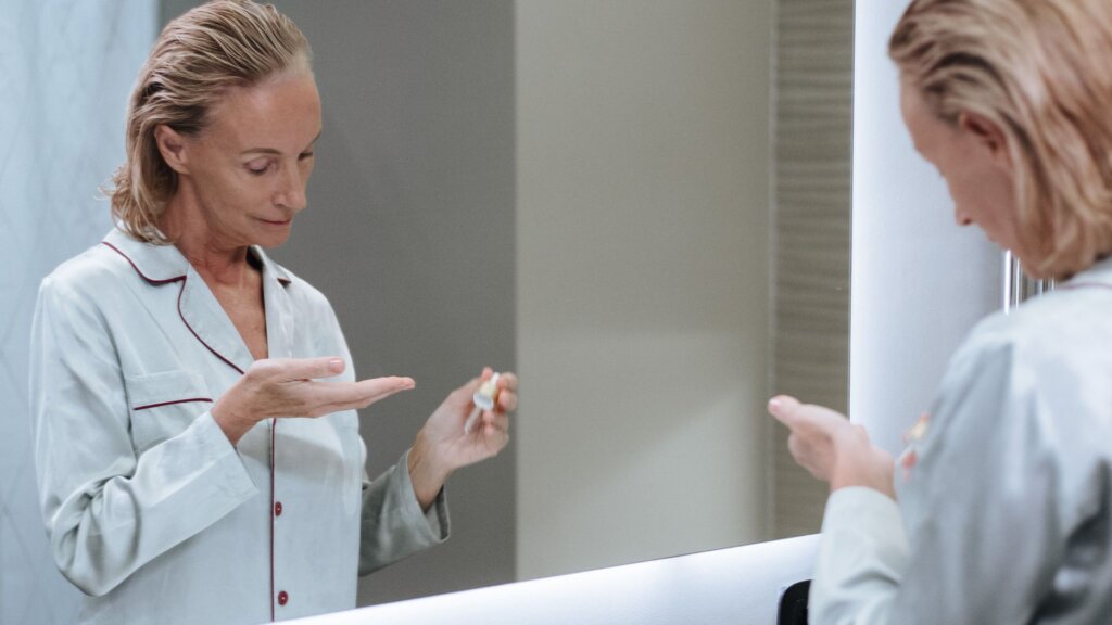 Woman dispensing skincare serum from a dropper into her hand at a bathroom mirror.
