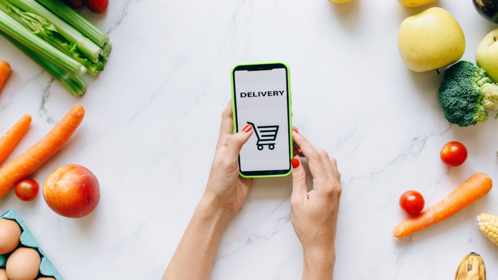 A person using a smartphone for grocery delivery surrounded by fresh fruits and vegetables.