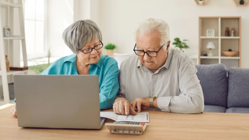 Senior couple reviewing financial documents on a laptop at a kitchen table, focused on their income calculations.