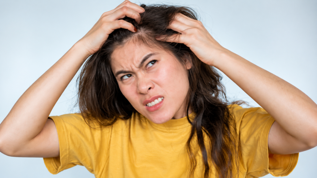 Woman in a yellow t-shirt scratching her scalp in frustration against a plain background.