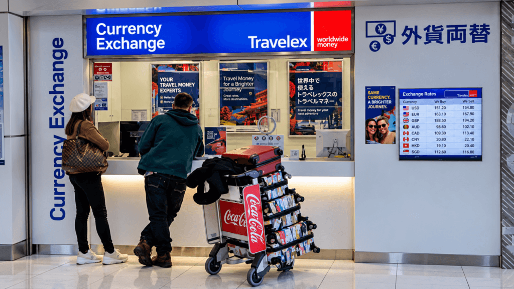 Two travelers standing at a Travelex Currency Exchange counter inside an airport next to a luggage cart.
