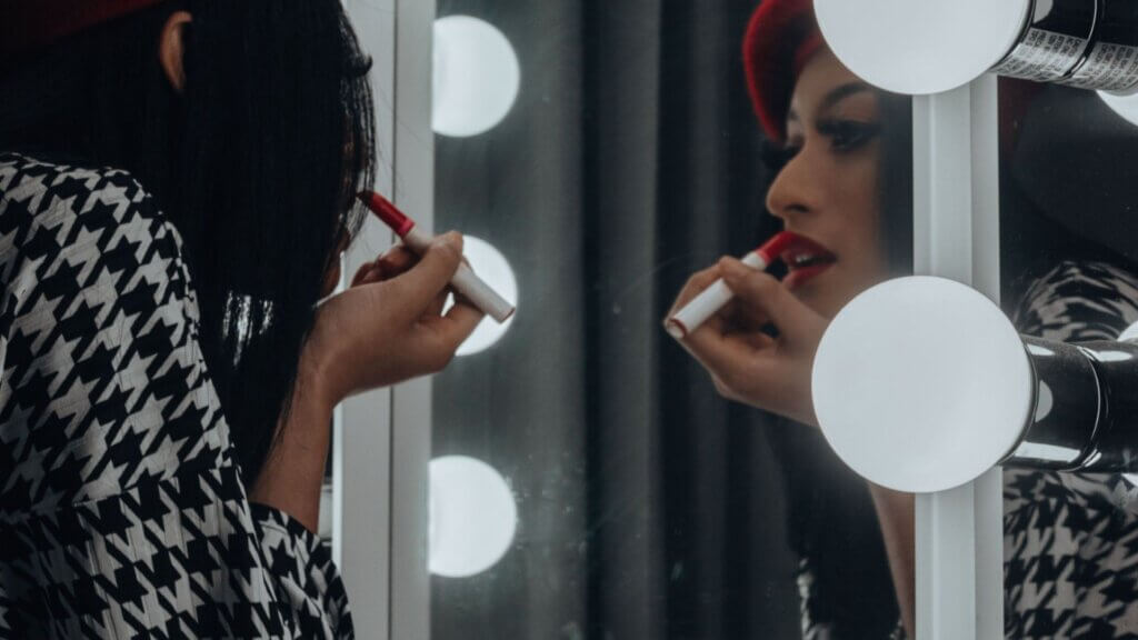 Person applying red lipstick while looking into a lighted makeup mirror.