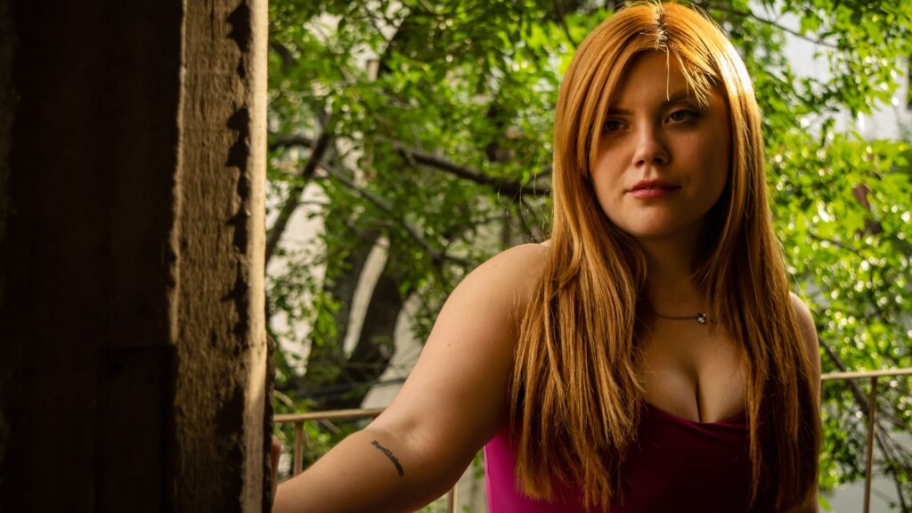 Woman with long red hair standing outdoors near trees and greenery.
