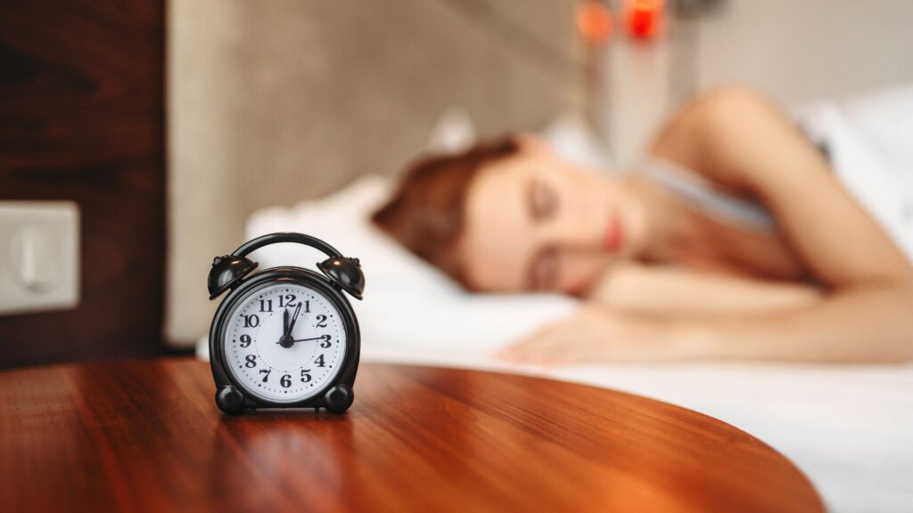 Alarm clock on a bedside table with a woman sleeping in the background.