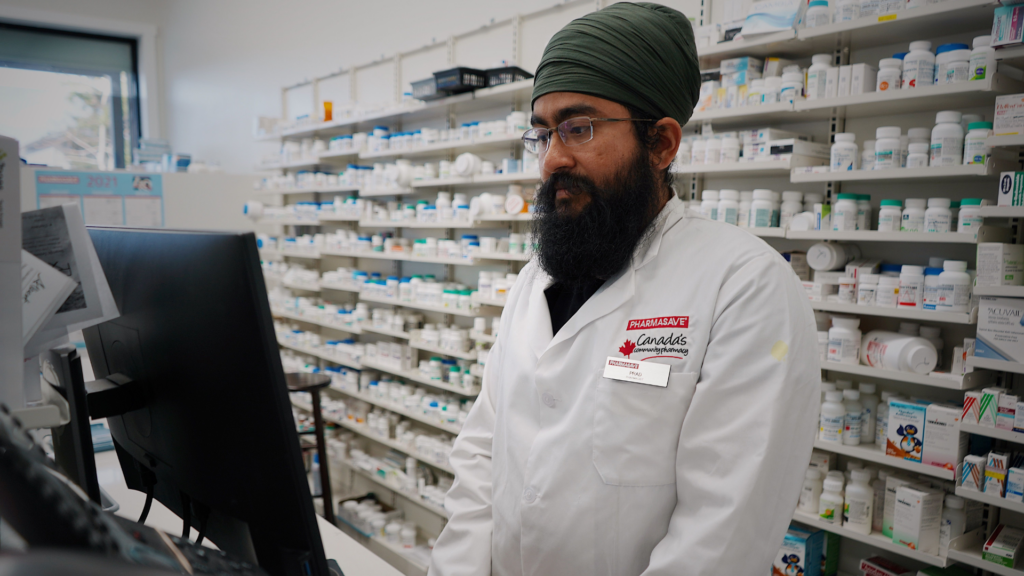 Pharmacist wearing a turban working at a computer in a pharmacy store with shelves filled with medicine.