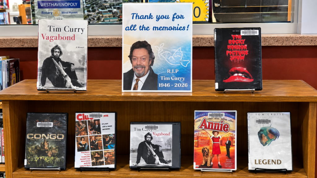 A library memorial display for actor Tim Curry featuring his memoir Vagabond and DVDs of his films like The Rocky Horror Picture Show, Clue, Congo, Annie, and Legend.