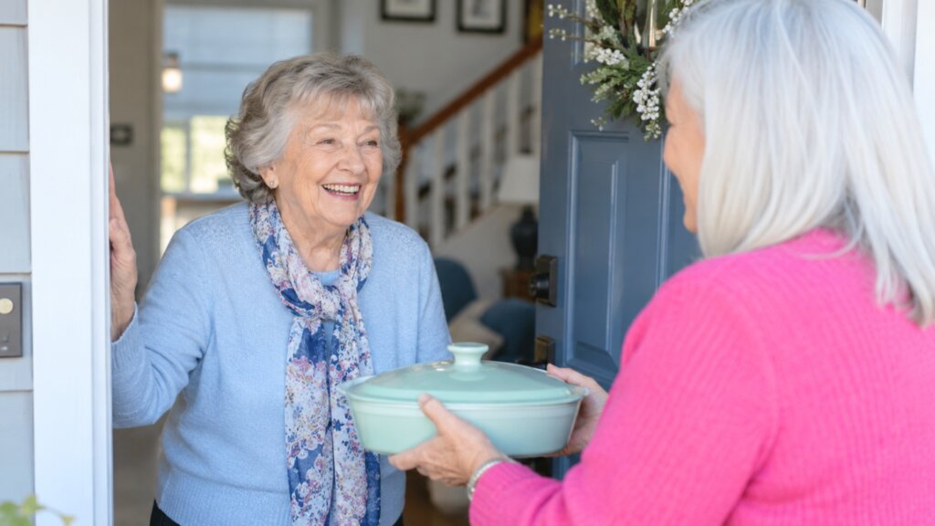 Smiling older woman welcomes another older woman at her front door as the visitor hands her a covered casserole dish. The friendly exchange suggests companionship and practical support between neighbors or friends.