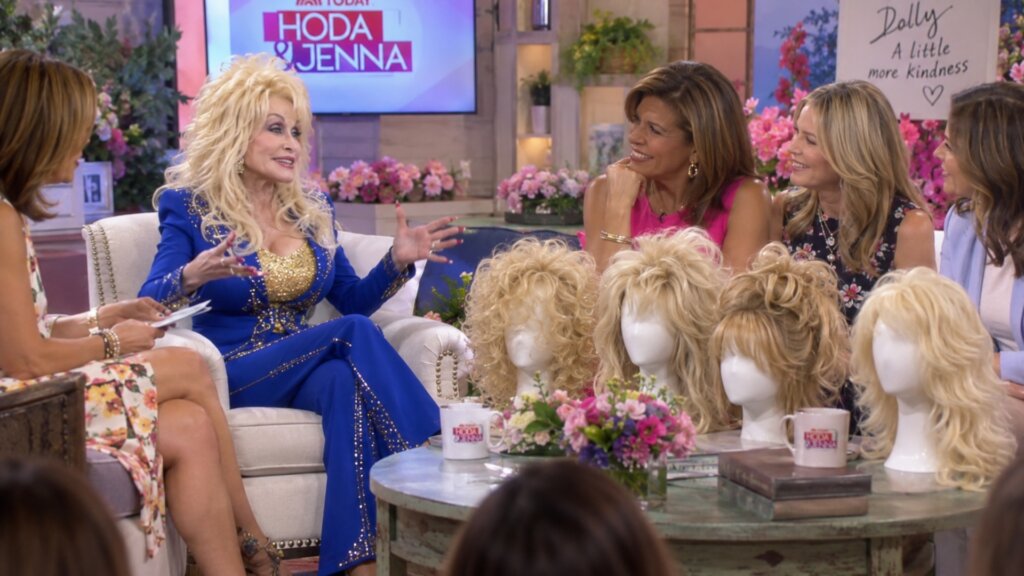 Dolly Parton in a show with several of her signature blonde wigs are displayed on mannequin heads.