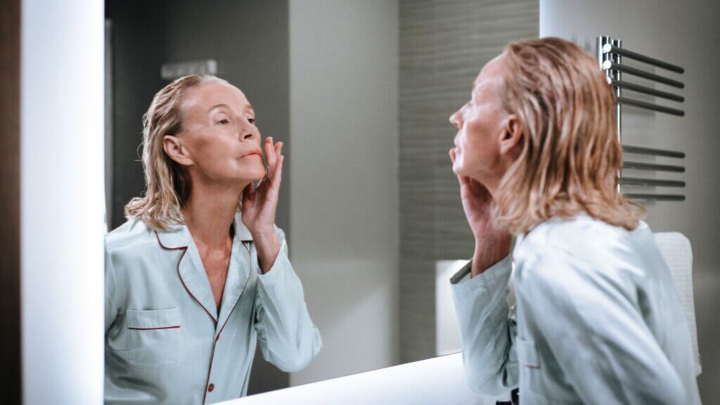 Older woman applying skincare to her face while looking in a bathroom mirror.