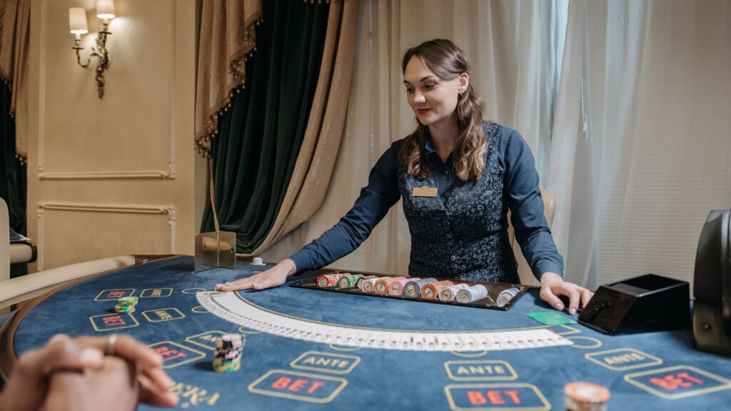 Casino dealer standing behind a gaming table with cards and poker chips.
