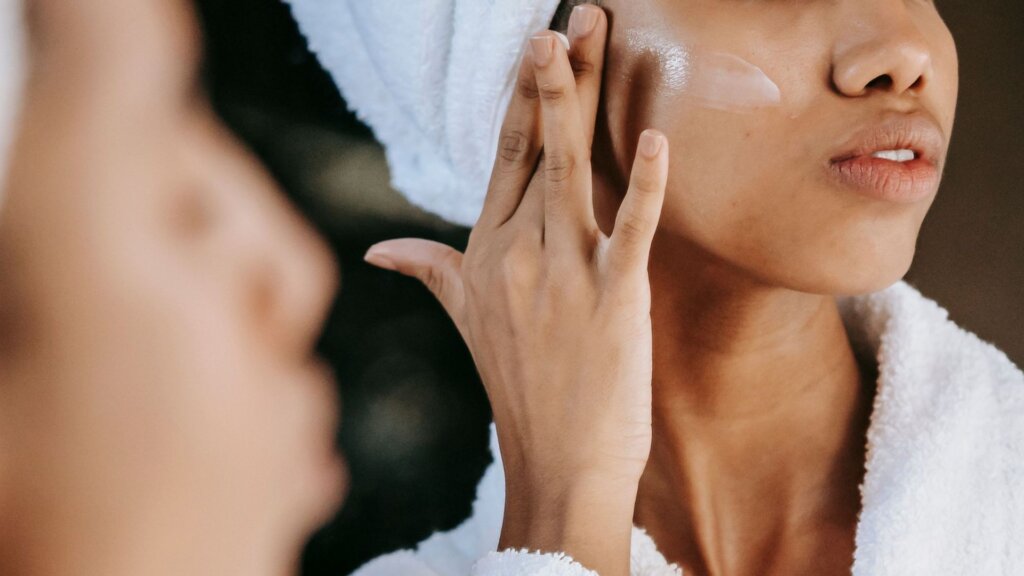 Woman applying skincare cream to her cheek while looking in a mirror.