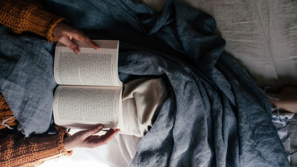 Person reading an open book while resting in bed under a gray blanket.