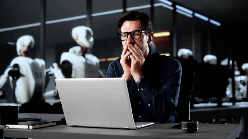 Stressed man looking at a laptop while humanoid robots work in the background.