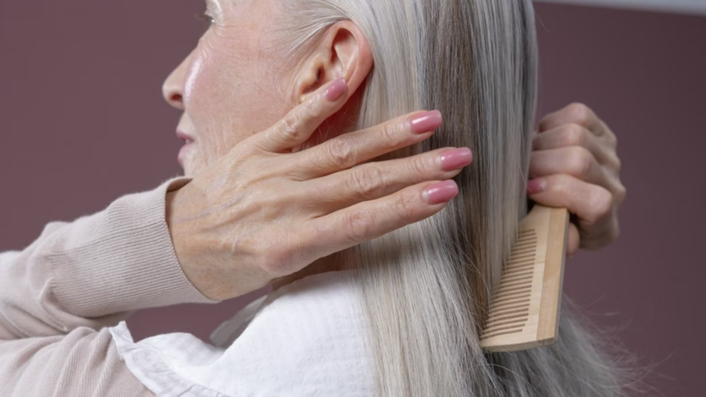 An older woman with long gray hair is shown from the side, brushing her hair with a wooden comb.