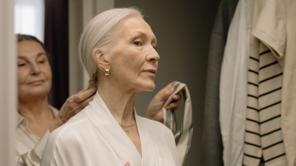 An older woman with white hair pulled back looks thoughtfully in a mirror as someone helps her adjust a white garment.
