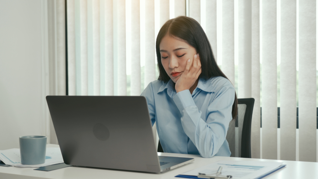 Exhausted woman sitting at a desk with a laptop, resting her chin on her hand.
