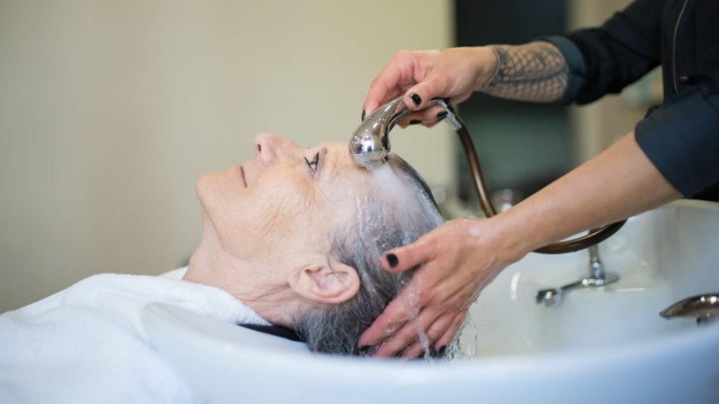 A senior woman is getting her hair washed in a salon sink, with the stylist holding a showerhead.