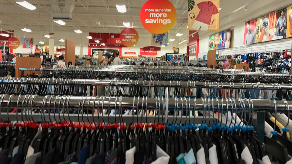 A wide interior shot of a T.J. Maxx store with rows of clothing racks, shoppers, and overhead promotional signs.