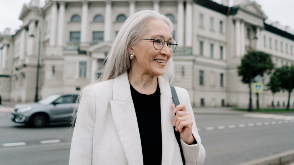 A smiling older woman with long gray hair and glasses stands outdoors in a city setting, wearing a light-colored coat.