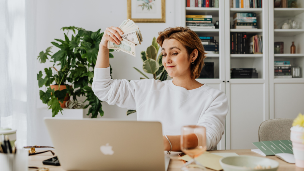 Smiling woman holding cash while working on a laptop in a cozy home office setting.