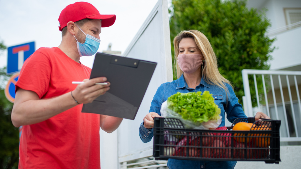 A delivery person in protective gear handing groceries to a customer at home.