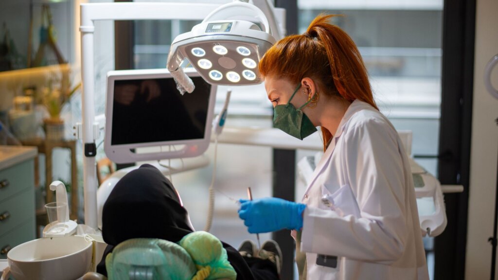 A dentist wearing a face mask and blue gloves examines a seated patient under a dental exam light.