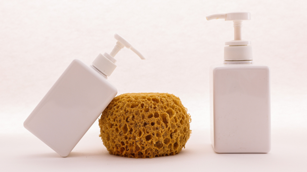 A minimalist setup shows two white pump bottles, one leaning, placed beside a round, natural sea sponge against a plain backdrop.