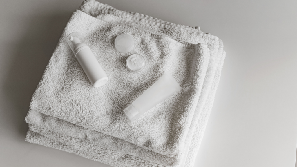 A top-down arrangement of skincare items, including a pump bottle, a small tub of cream, and a squeeze tube, resting on a folded white towel.