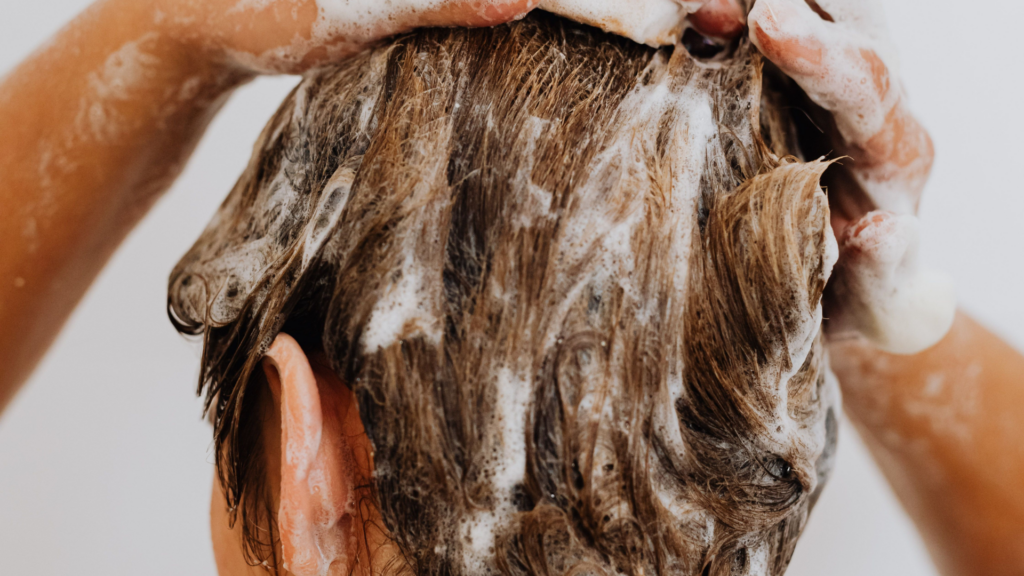 A view from behind shows a person with short brown hair massaging sudsy white shampoo through their hair in the shower.