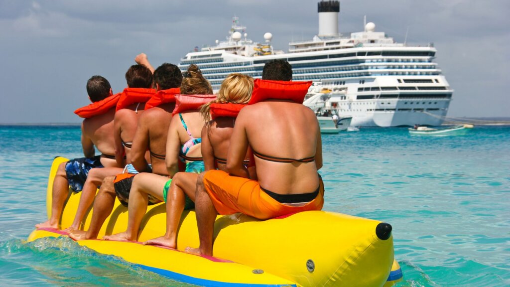 People ride a yellow inflatable banana boat across turquoise water, wearing red life jackets, with a cruise ship in front of them.
