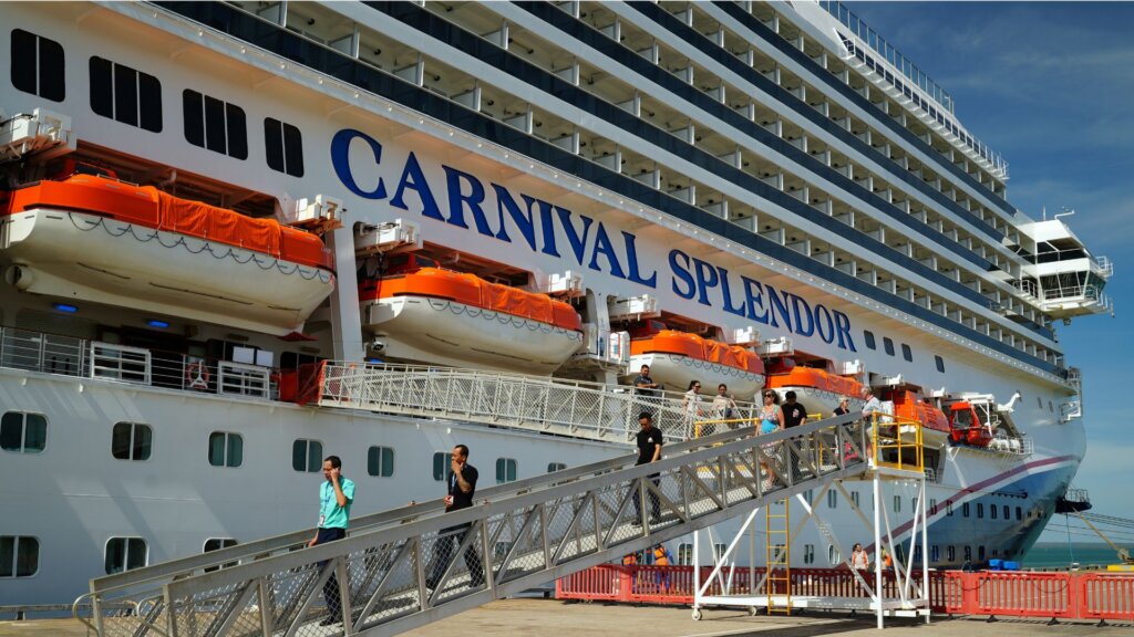 Carnival Splendor cruise ship docked at port, with orange lifeboats, passengers on a gangway, and blue sky.