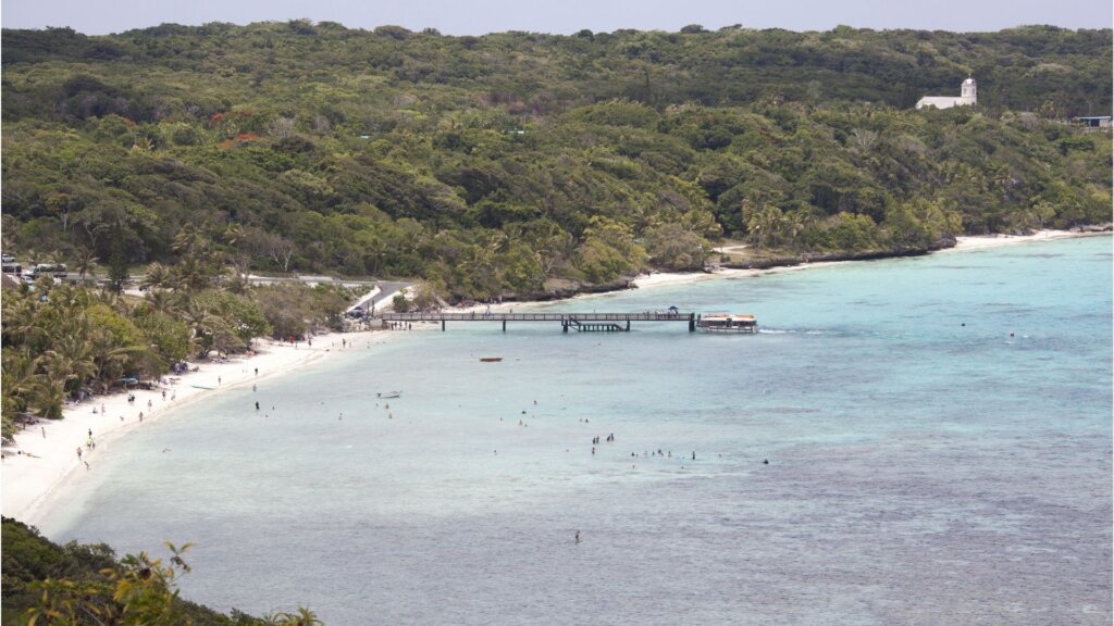 Aerial view of a beach on Lifou Island and a turquoise bay, with swimmers, a pier, and forested hills.