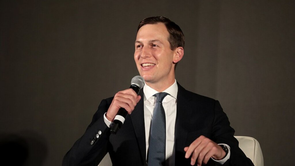 Jared Kushner smiling and holding a microphone while seated in a white chair.