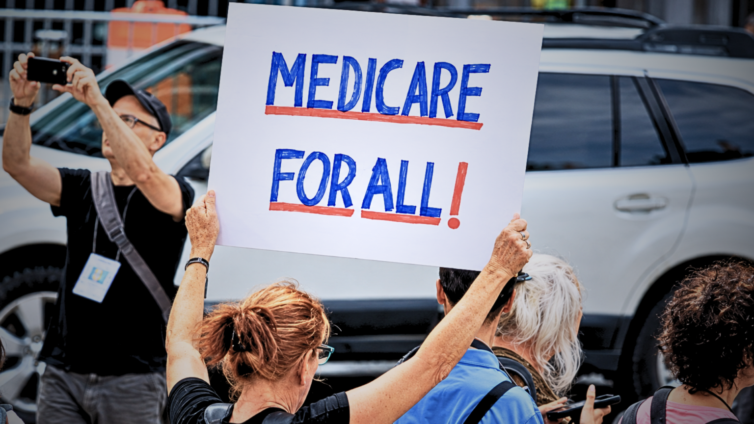 Demonstrators holding up a sign that reads "MEDICARE FOR ALL!" in front of a white SUV.