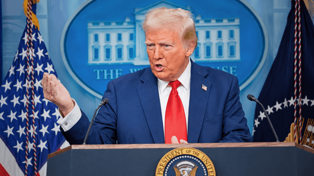Donald Trump standing behind a podium with the Presidential seal, speaking and gesturing with one hand in front of the White House press backdrop.