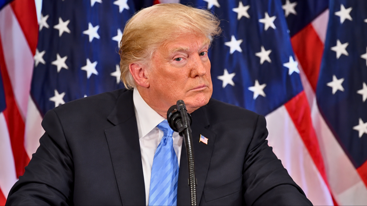 Donald Trump sitting in front of several American flags, looking forward with a serious expression.