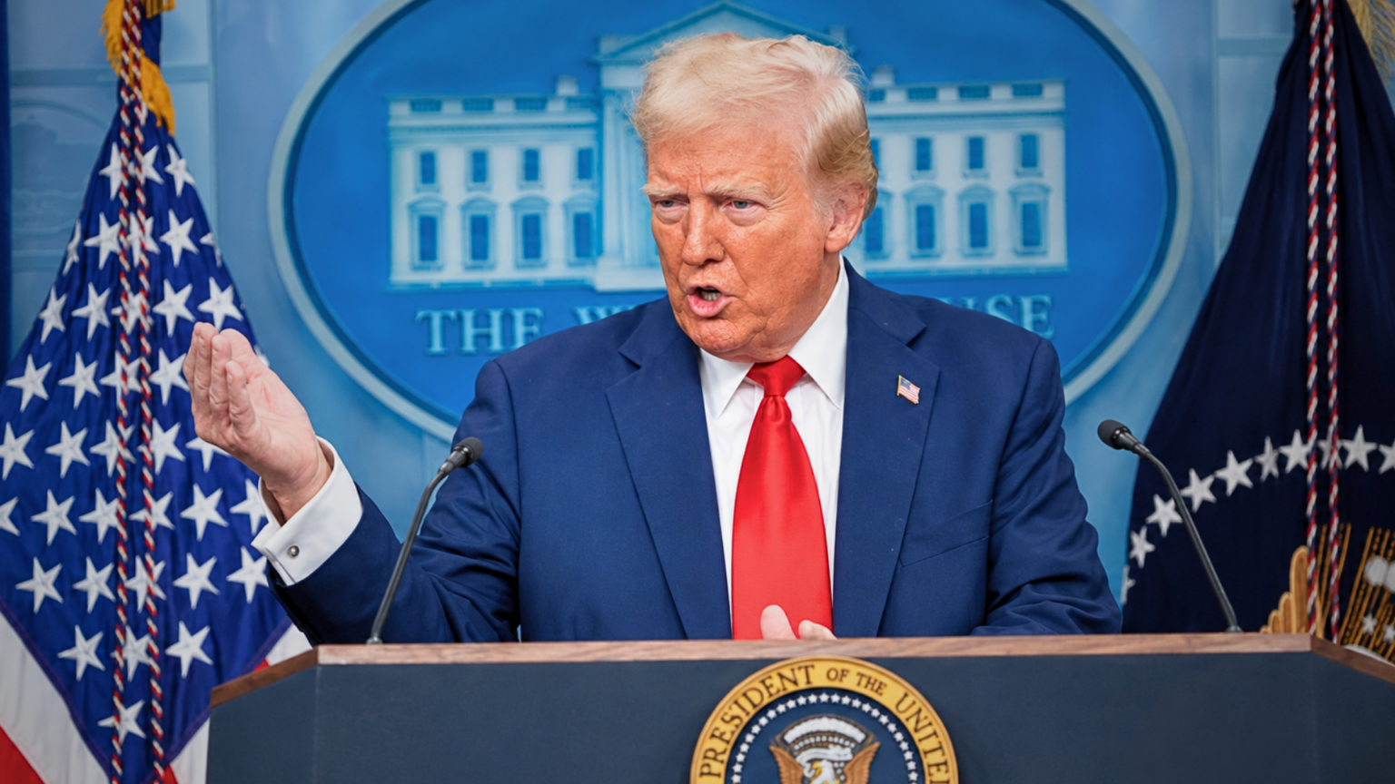 Donald Trump standing behind a podium with the Presidential seal, speaking and gesturing with one hand in front of the White House press backdrop.