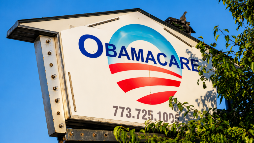 An outdoor sign displaying the word "OBAMACARE" with a red, white, and blue logo and a phone number against a clear blue sky.